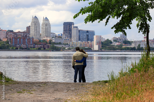 A young man and girl are standing and hugging on a cloudy morning on the background of Dnieper and skyscrapers. Spring, summer cityscape, big city. Panorama of Dnepropetrovsk, Dnipro, Ukraine.