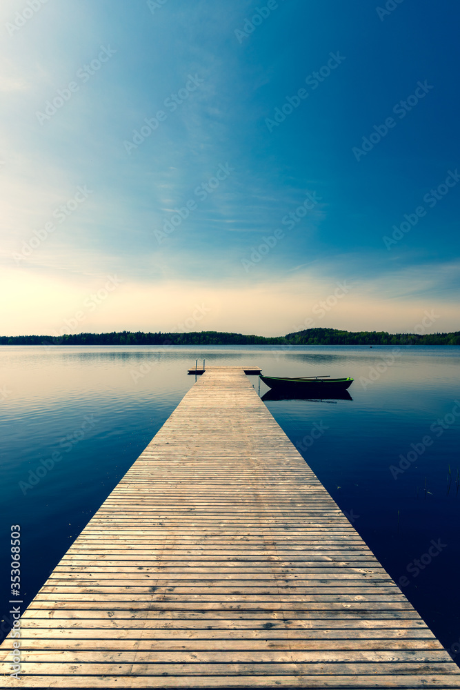 A wooden boat with oars moored to a pier on a lake in spring. The pier from the planks leaves with the prospect towards the horizon. The sun in the blue sky is approaching sunset. Latvia