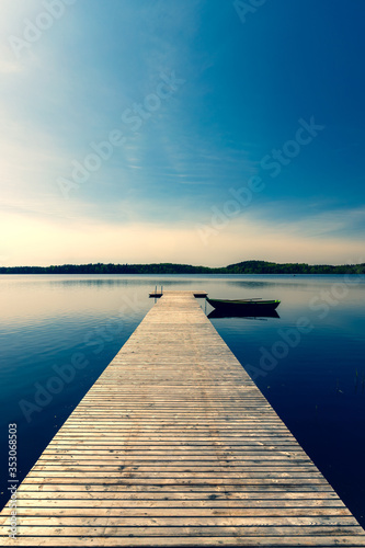 Fototapeta Naklejka Na Ścianę i Meble -  A wooden boat with oars moored to a pier on a lake in spring. The pier from the planks leaves with the prospect towards the horizon. The sun in the blue sky is approaching sunset. Latvia