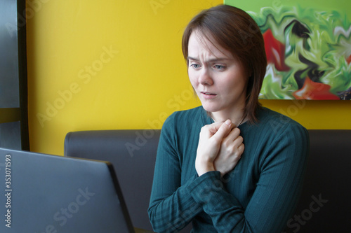 A girl sits, works at a laptop at a table in a cafe on a brown sofa against a yellow wall. On the face is an expression of compassion, grief, tension. The steering wheels are folded and pressed to the