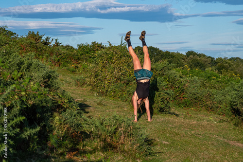 Young girl in a hat doing a handstand on the green hill. Hiking and sport lifestyle concept.