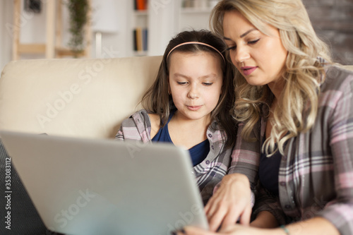 Mother helping little daughter to type on laptop keyboard sitting on the ouch in living room.