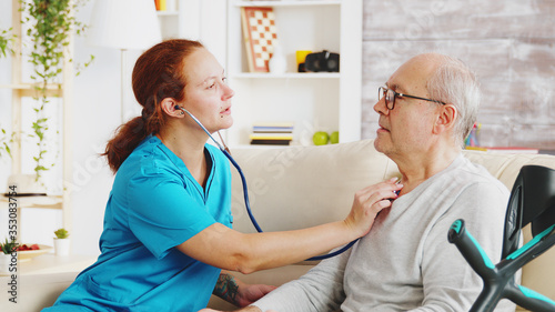 Caucasian female nurse listening to elderly retired man heartbeat in bright and cozy nursing home. Social worker and caregiver