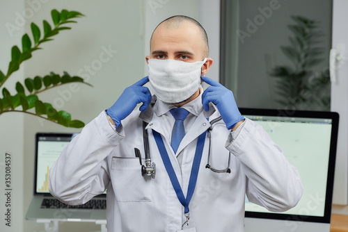 A caucasian doctor correcting a protective face mask to avoid the spread coronavirus  COVID-19  in his office. A physician with a beard preparing to examine a patient.