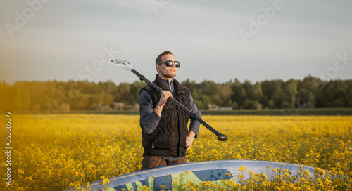 A young man with a paddle and sup Board in a rapeseed field