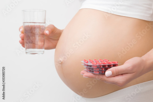 Young woman hands holding water glass and blister packs of red pills for improve of iron level in blood. Receiving vitamins in pregnancy time. Side view. Close up. Light gray background.