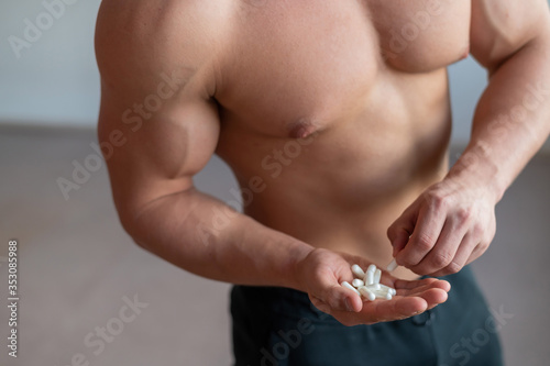 Faceless bodybuilder drinks growth hormone capsules. A close-up of a muscular man with a naked torso holds a handful of vitamins and fitness supplements.