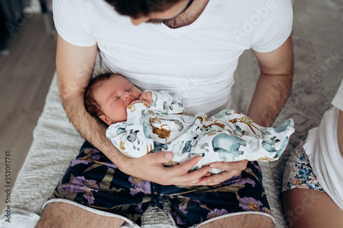 Candid real family moment. Soft focus. Young dark hair father holding his newborn baby son sitting in a bedroom. Boy is wearing body with watercolor animal print. Father's day. Boy is sleeping