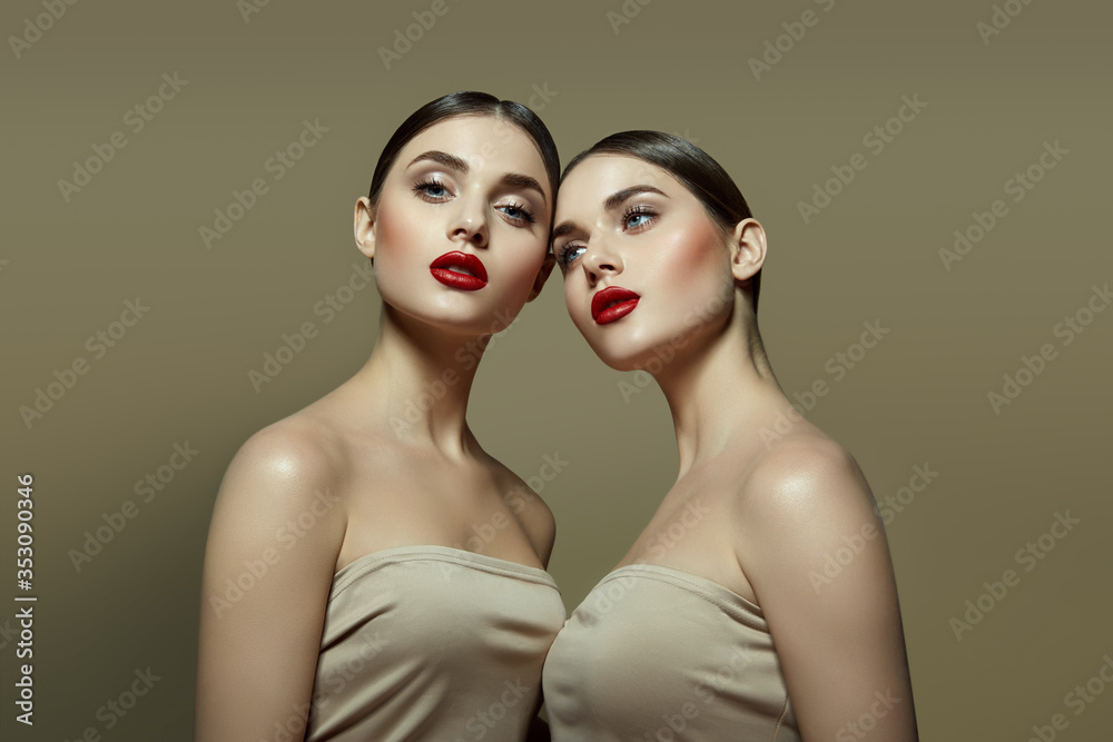 Two beautiful young girls with collected dark hair with perfect skin and bright red lips in beige tops on a beige background in the studio.