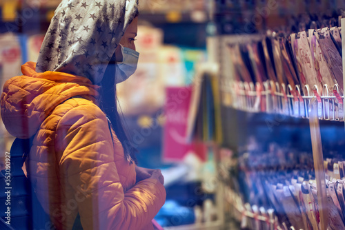 A girl in a medical mask in an orange jacket., hood and briefcase, chooses an item in the store. covid-19. the background is indistinct. shopping