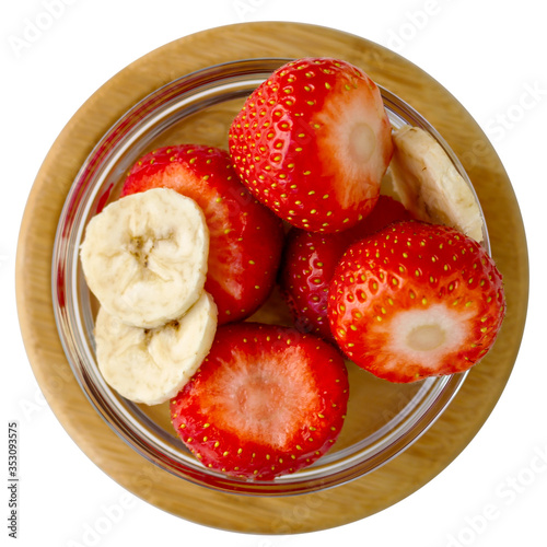 Whole fresh strawberries and banana slices in a round glass bowl on a wooden stand isolated on white background