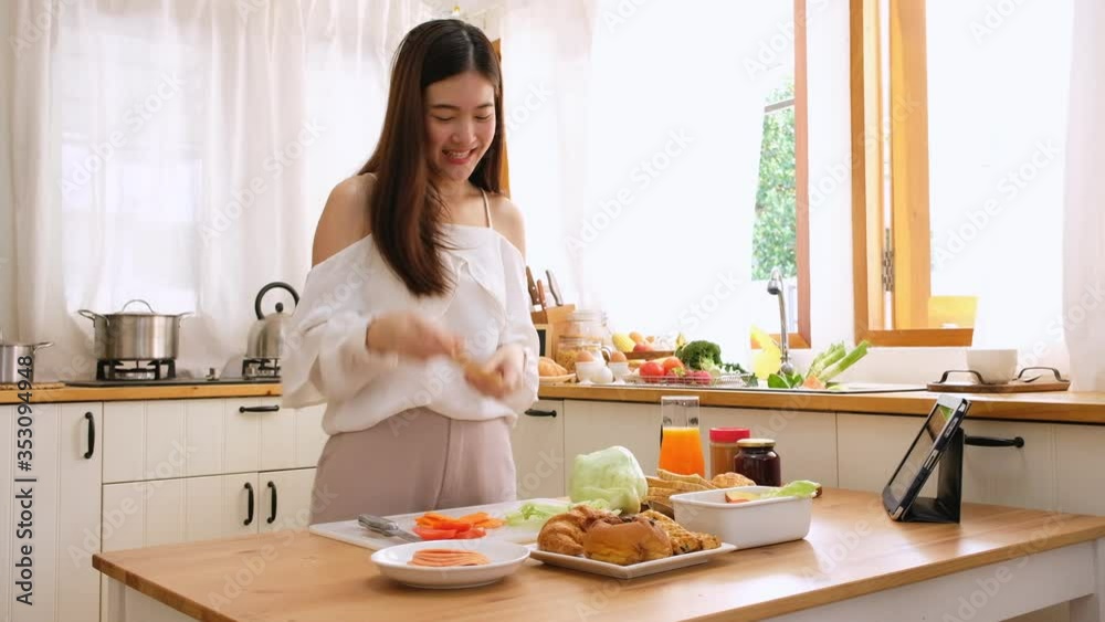Cute asian woman with brightly smile doing sandwich and chatting to friends with tablet in the kitchen Cute asian woman with brightly smile doing sandwich and chatting to friends with tablet in the kitchen