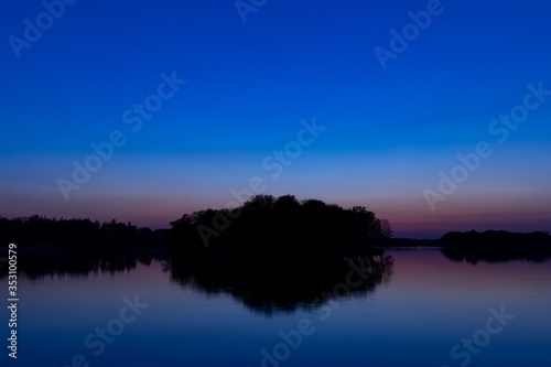 Fototapeta Naklejka Na Ścianę i Meble -  twilight landscape after sunset scenic view horizon silhouette of land with peaceful waters and reflection surface island and trees center of composition, dark blue sky lighting