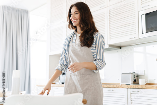 Image of pleased brunette woman wearing apron smiling while cooking pie