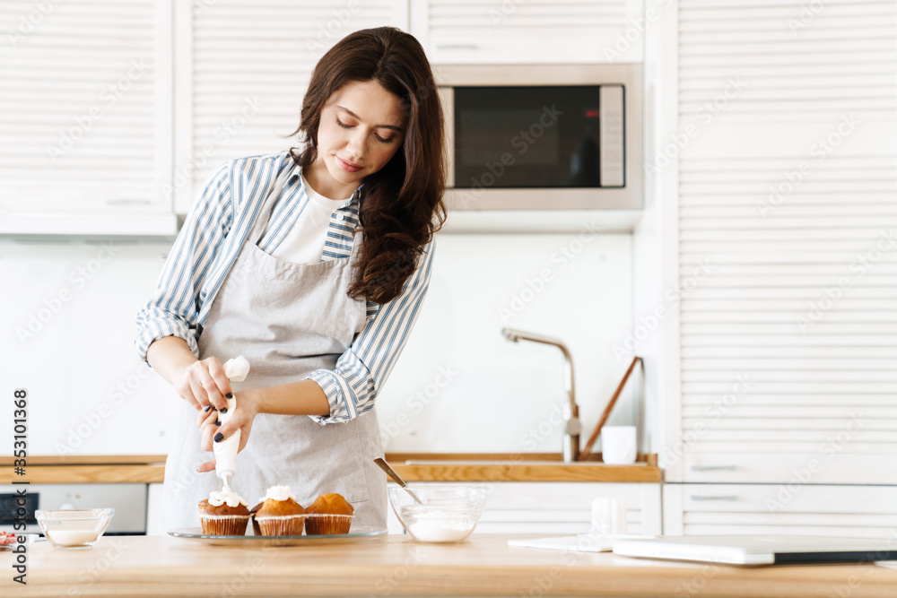 Image of pleased brunette woman cooking muffins with cream