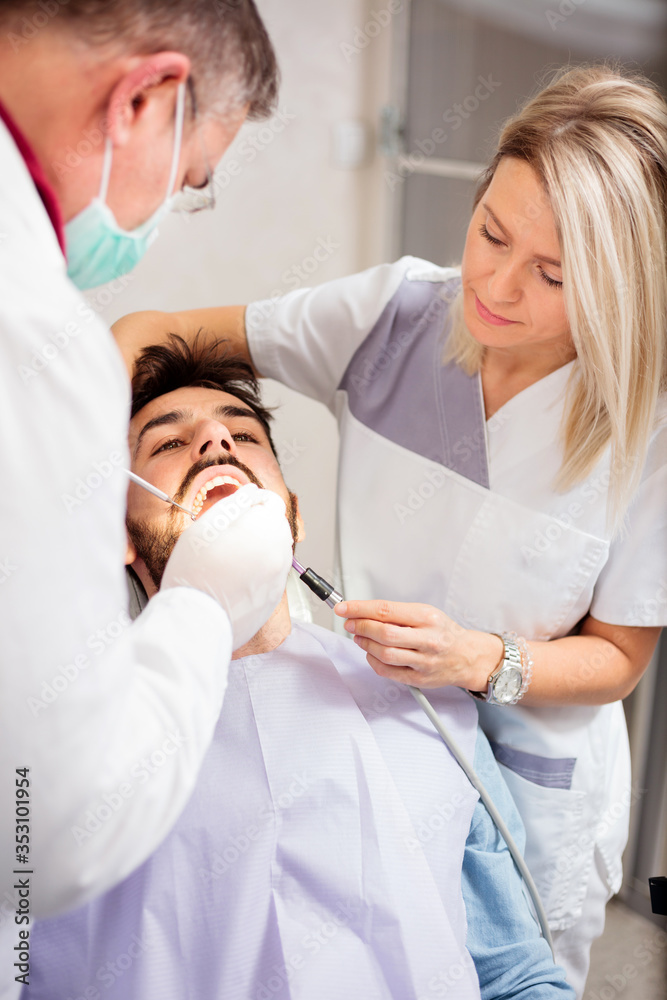 Young Caucasian man having his teeth examined in dental clinic by a mature male dentist with the help of a young female assistant