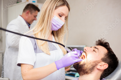Happy young female Caucasian dentist drilling out dental cavity and preparing the tooth for filling. Young man having his teeth checked in a dental clinic