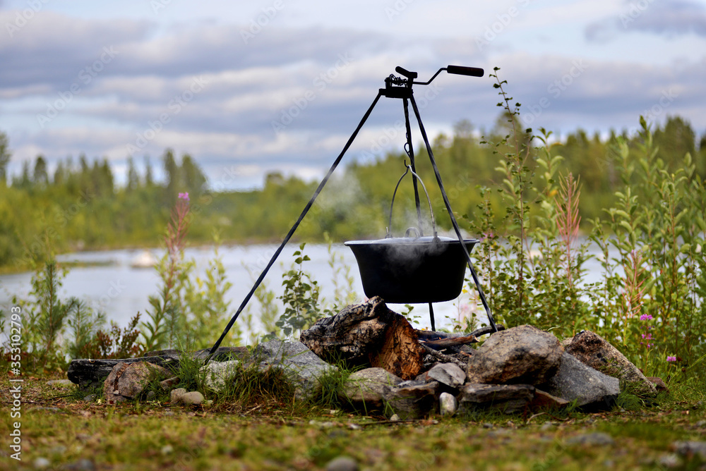 Hanging pot on a tripod in a fireplace lined with stones with forest, sky and river in the background Hanging pot on a tripod in a fireplace lined with stones with forest, sky and river in the background