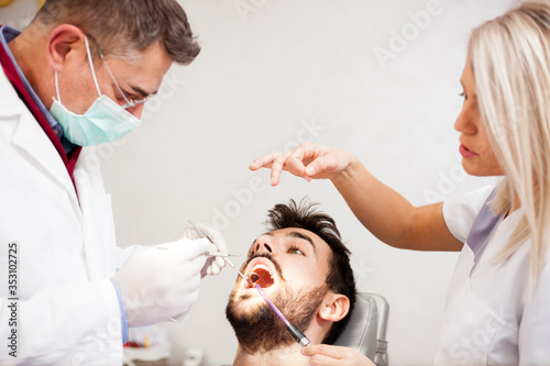 Young Caucasian man having his teeth examined in dental clinic by a mature male dentist with the help of a young female assistant