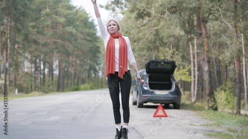 Desperate blond woman hitchhiking on suburban road. Wide shot portrait of stressed Caucasian female driver having car breakage on countryside highway. Problem, accident, lifestyle, travelling.