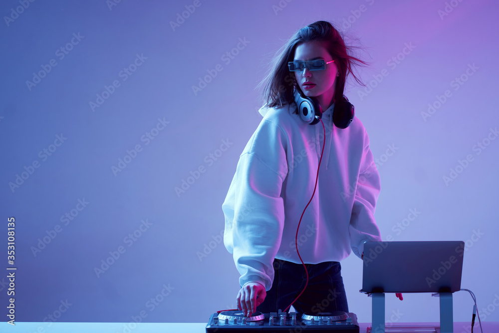 Cool young girl DJ mixes music on a mixing console and laptop, in stylish clothes, glasses on a neon background.