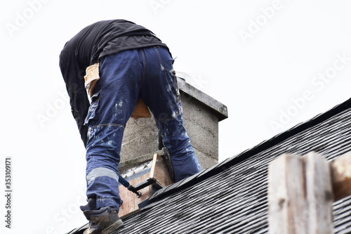 Roofer construction worker repairing chimney on grey slate shingles roof of domestic house, sky background with copy space.
