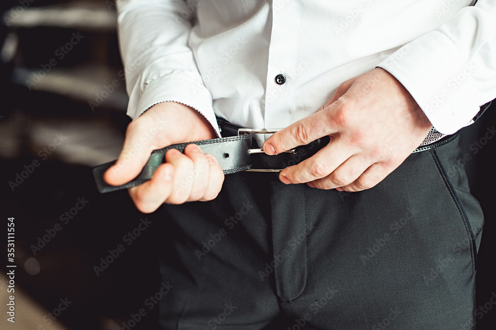 Groom in black pants and a white shirt fastens his leather belt. grooms mourning. Wedding details. Groom in black pants and a white shirt fastens his leather belt. grooms mourning. Wedding details.