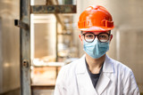 Tecnico specialista in camice bianco e caschetto protettivo arancione indossa una mascherina chirurgica e degli occhiali tecnici , in contesto industriale.