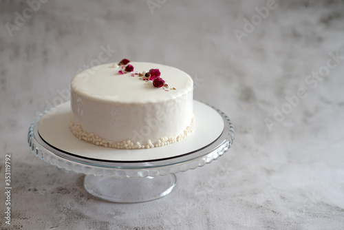 Сake stands on a round transparent glass stand on a gray texture background. Siimple white mousse cake decorated with dried rose buds on smooth glaze.