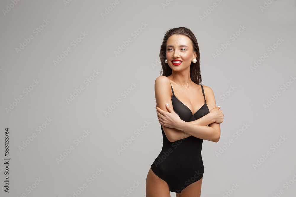 Obraz premium Beautiful young caucasian female with long dark hair, nice make up, red lips in black swimsuit smiles