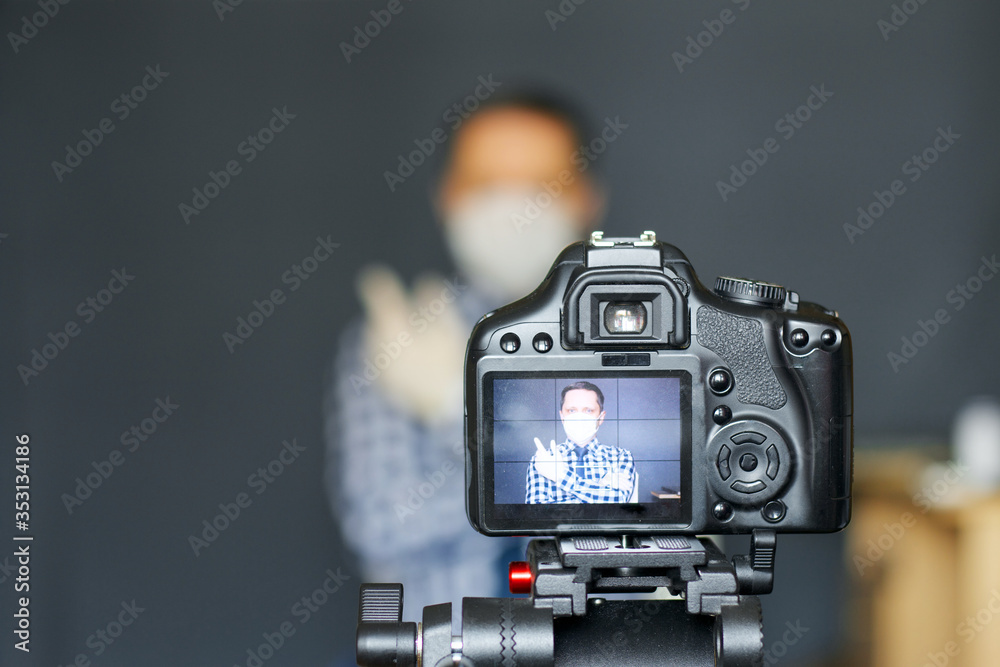 A blogger in a protective mask takes pictures of himself on camera. Focus is on his camera on a tripod and the image on it. Everything else is blurry.