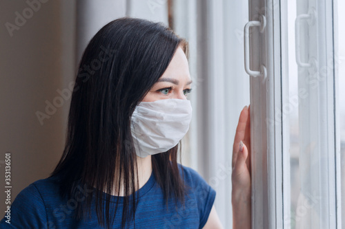 Sad quarantined woman with covid-19 wearing a face mask looking at window