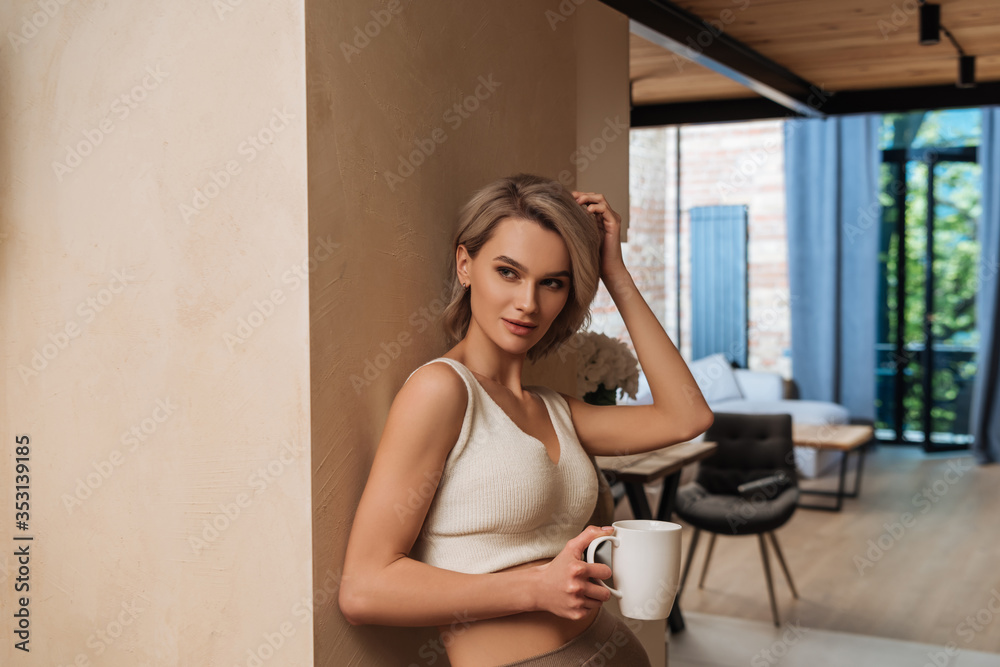 beautiful woman holding cup of tea and touching hair while standing by wall and looking away at home