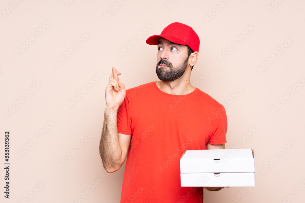 Young man holding a pizza over isolated background with fingers crossing and wishing the best Young man holding a pizza over isolated background with fingers crossing and wishing the best
