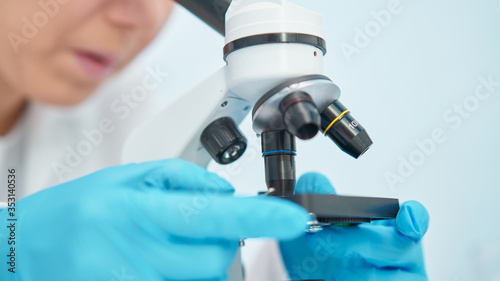 Close-up of female lab technician in blue rubber gloves looking through microscope.