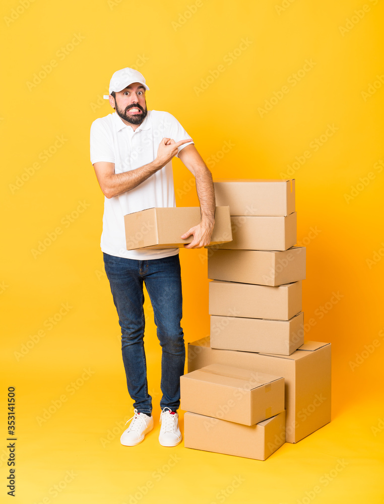 Full-length shot of delivery man among boxes over isolated yellow background frightened and pointing to the side Full-length shot of delivery man among boxes over isolated yellow background frightened and pointing to the side