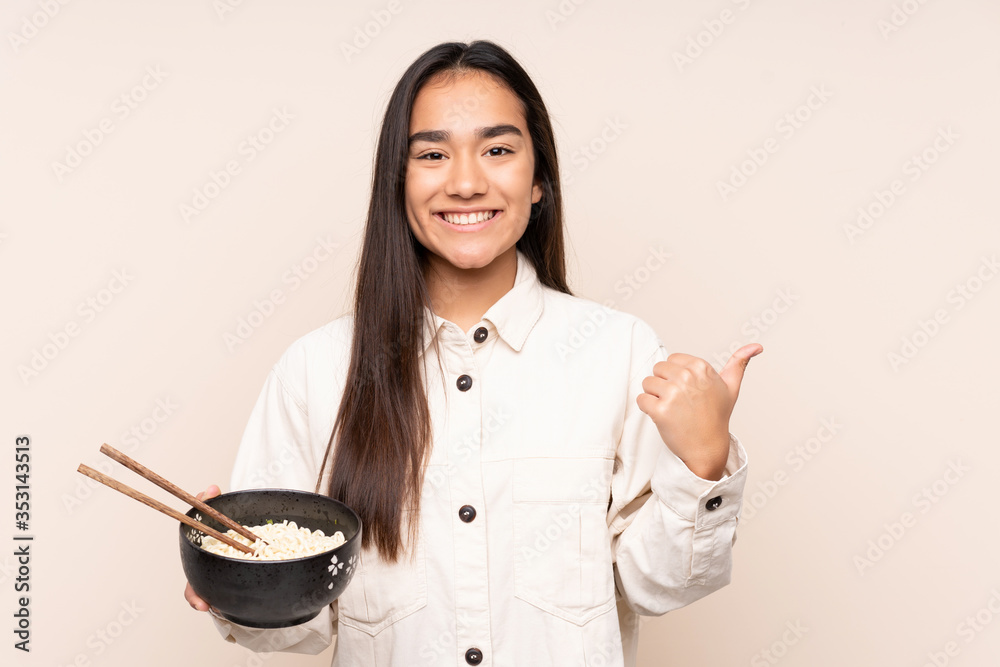 Young Indian woman isolated on beige background pointing to the side to present a product while holding a bowl of noodles with chopsticks Young Indian woman isolated on beige background pointing to the side to present a product while holding a bowl of noodles with chopsticks