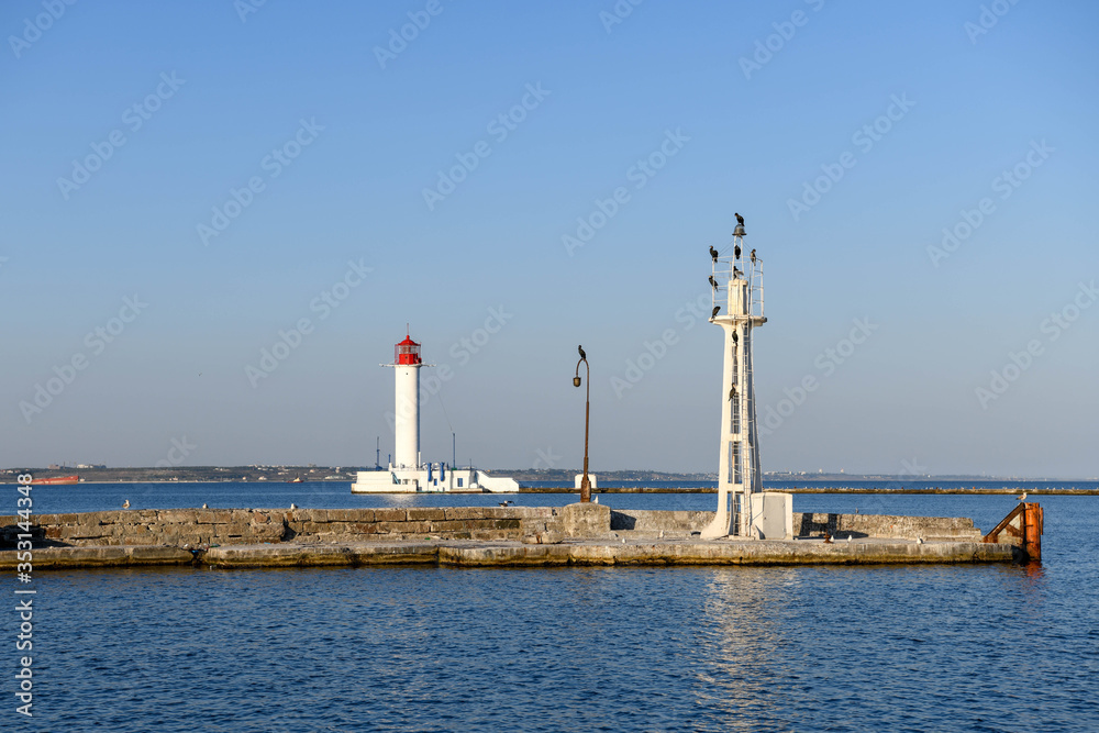 Fototapeta premium Birds are perched on a tower and the Odessa lighthouse is in the background illuminated by the setting sun.