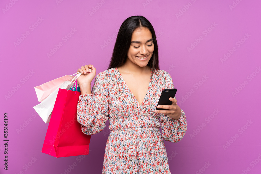 Young Indian woman isolated on purple background holding shopping bags and writing a message with her cell phone to a friend Young Indian woman isolated on purple background holding shopping bags and writing a message with her cell phone to a friend