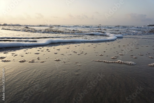 Rolling waves on the seashore with froth and bubbles on the wet sand during dusk or dawn taken from a low angle with scattered clouds over horizon.