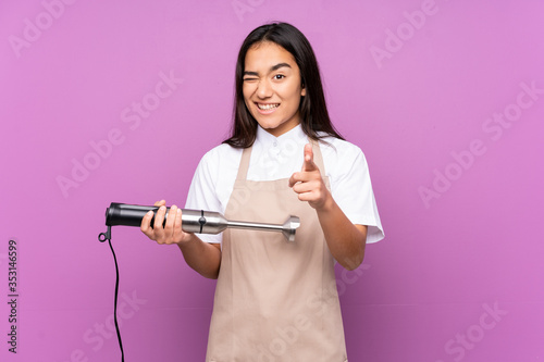 Indian woman using hand blender isolated on purple background points finger at you