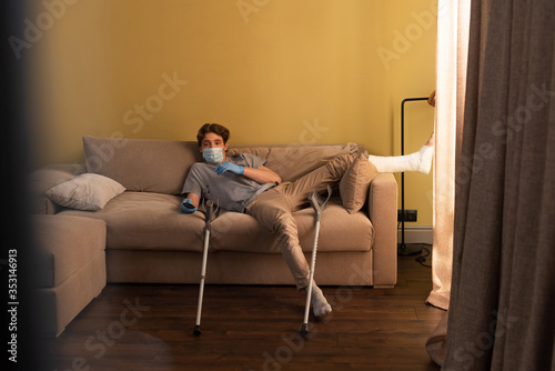 Selective focus of man in medical mask, latex gloves and plaster bandage on leg watching tv at home