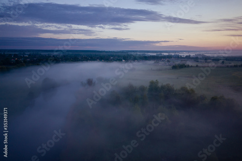 The river and meadow are covered with fog until sunrise. Pre-dawn, hazy landscape. Trees peek through the fog.