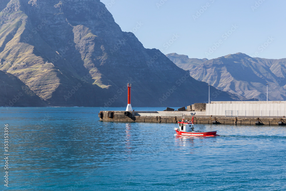 small red and white fishing boat sails through the port of Galdar with high mountains in the background