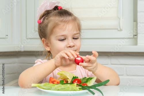 little girl sits at a table and eats vegetables. red radish in hand
