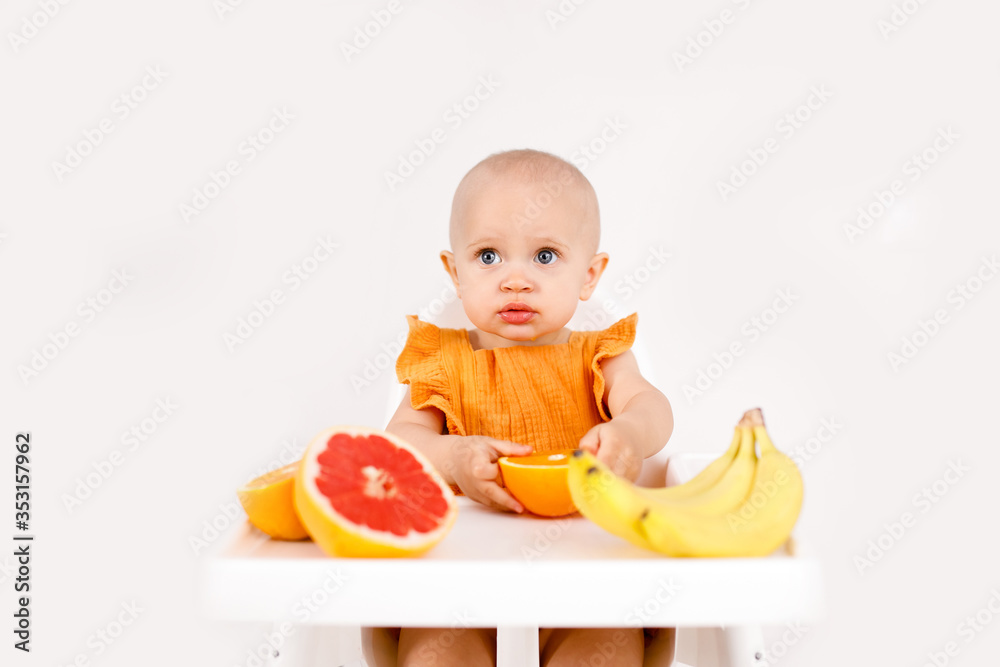 Happy baby sitting in high chair eating fruits in a white kitchen. Healthy nutrition for kids. Children eat oranges and grapefruits Happy baby sitting in high chair eating fruits in a white kitchen. Healthy nutrition for kids. Children eat oranges and grapefruits