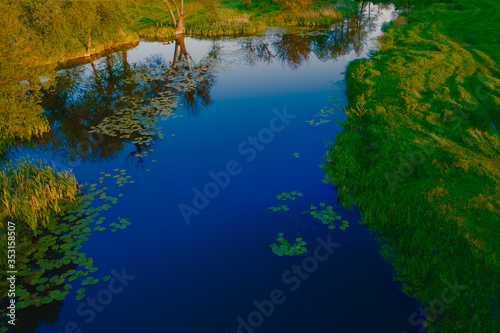 Evening river landscape. Pure blue calm water surface among the luscious green lawn outside the city.  © Andrii Chagovets