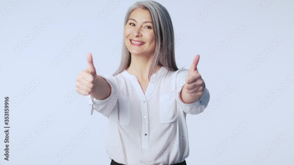 A smiling good-looking old mature woman with long gray hair wearing formal business clothes is doing a thumb up gesture with both hands standing isolated over white background in studio A smiling good-looking old mature woman with long gray hair wearing formal business clothes is doing a thumb up gesture with both hands standing isolated over white background in studio