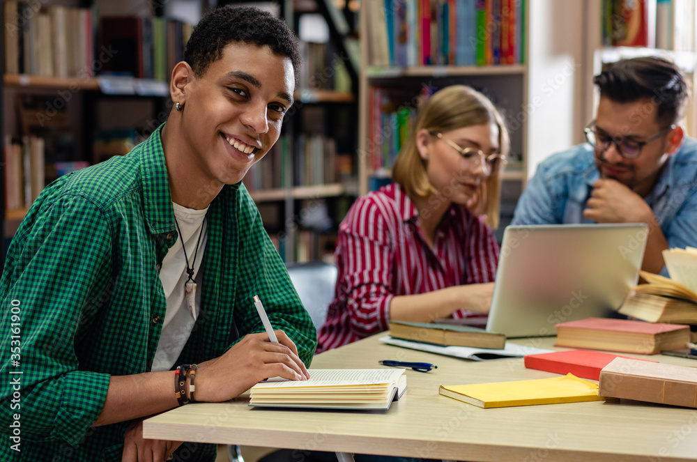 Portrait of young african american student sitting in library looking at camera, with his friends in background. Portrait of young african american student sitting in library looking at camera, with his friends in background.