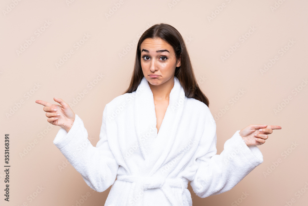 Young girl in a bathrobe over isolated background pointing to the laterals having doubts Young girl in a bathrobe over isolated background pointing to the laterals having doubts